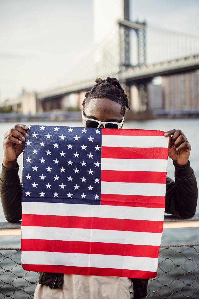 services-03 African American male in sunglasses with American Flag bandana in hands leaning on waterfront railing against blurred modern city