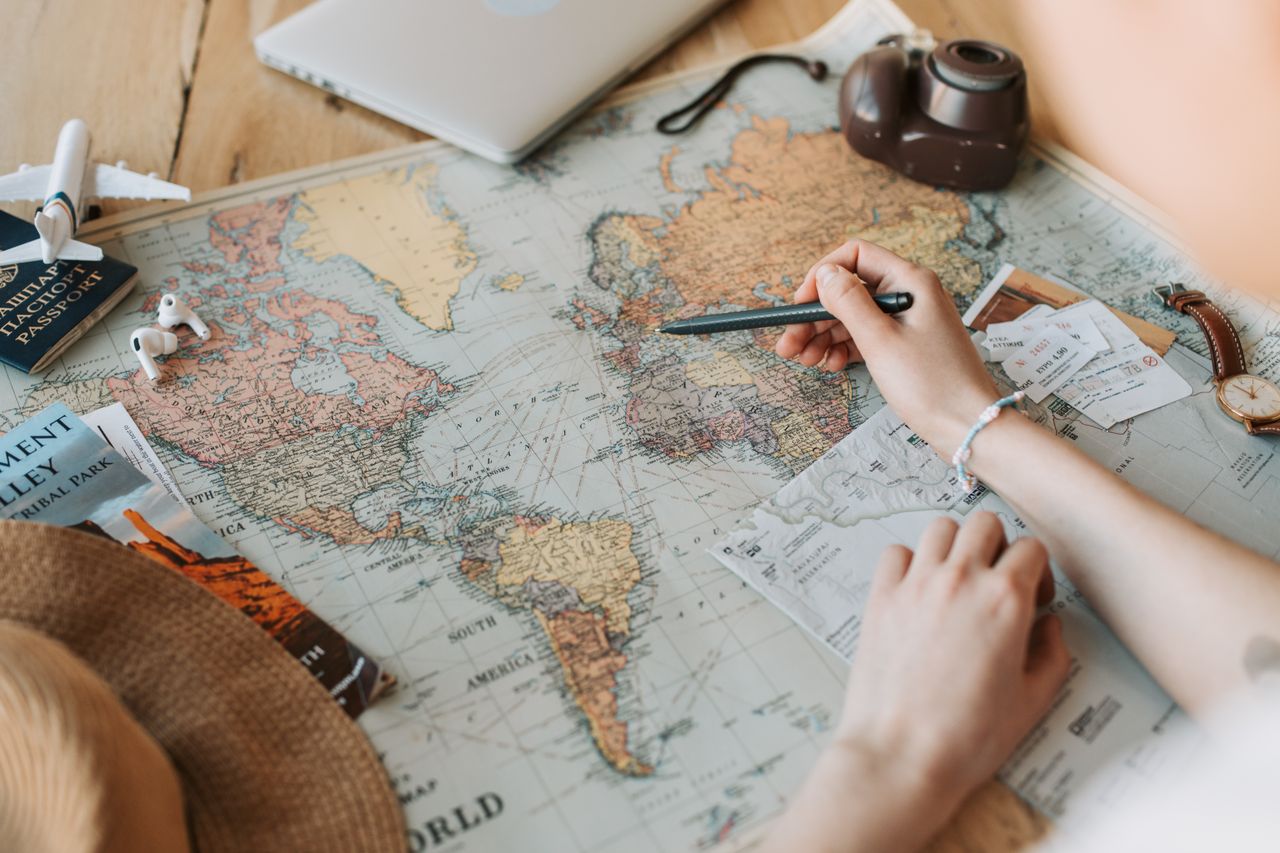 services-02 Hands marking destinations on a world map surrounded by travel essentials like passport and camera.